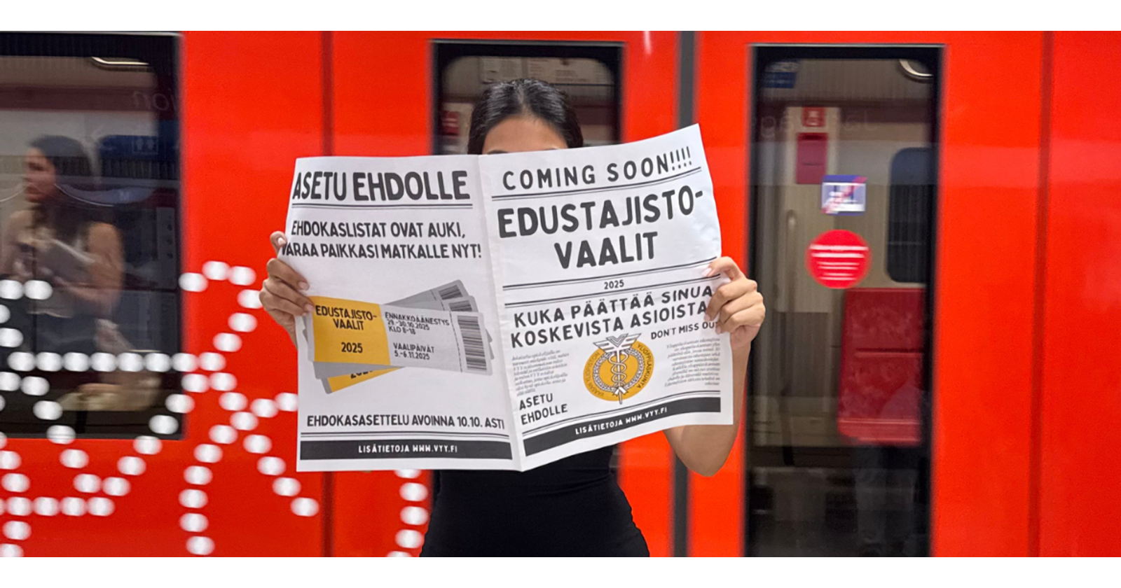 Nainen seisoo metroasemalla pidellen VYY-aiheista sanomalehteä auki kasvojensa edessä. Taustalla kulkee punainen metro.