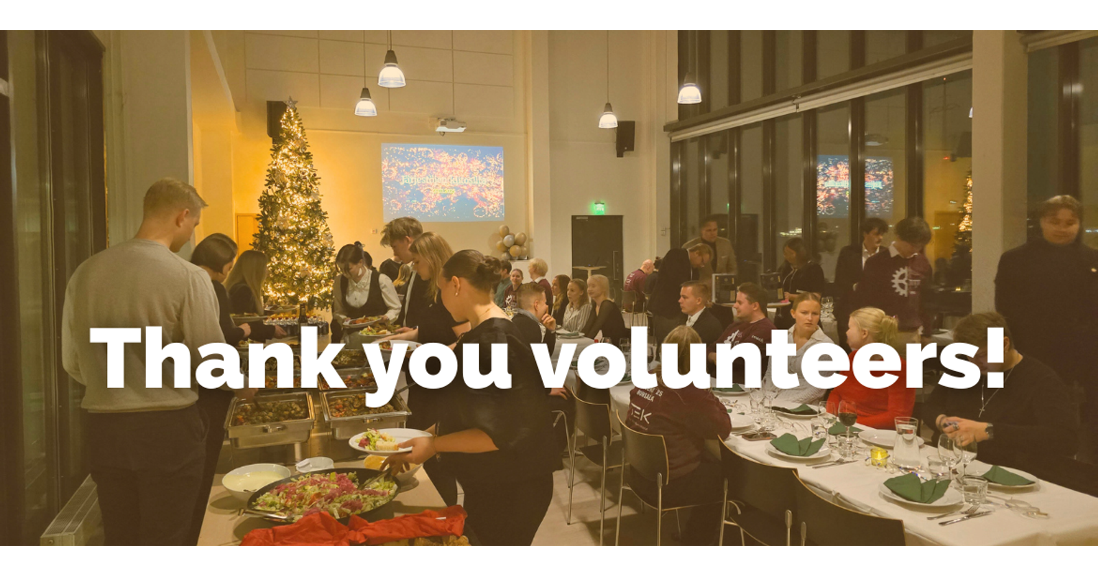 Photograph shows people at a dinner celebration, some are sitting and others are getting food from the buffet. There's a Christmas tree in the background. Above the photo there's text Thank you volunteers!