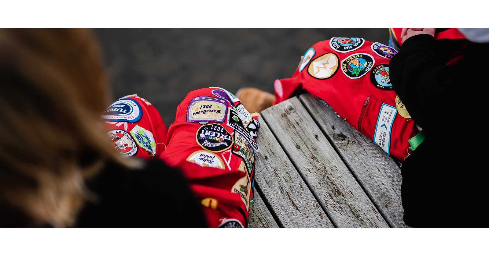 Two people are sitting on a dock wearing red overalls and dark tops. There is dark water in the background. The image is taken from the shoulder level of the people in the picture towards the overalls and the dock.