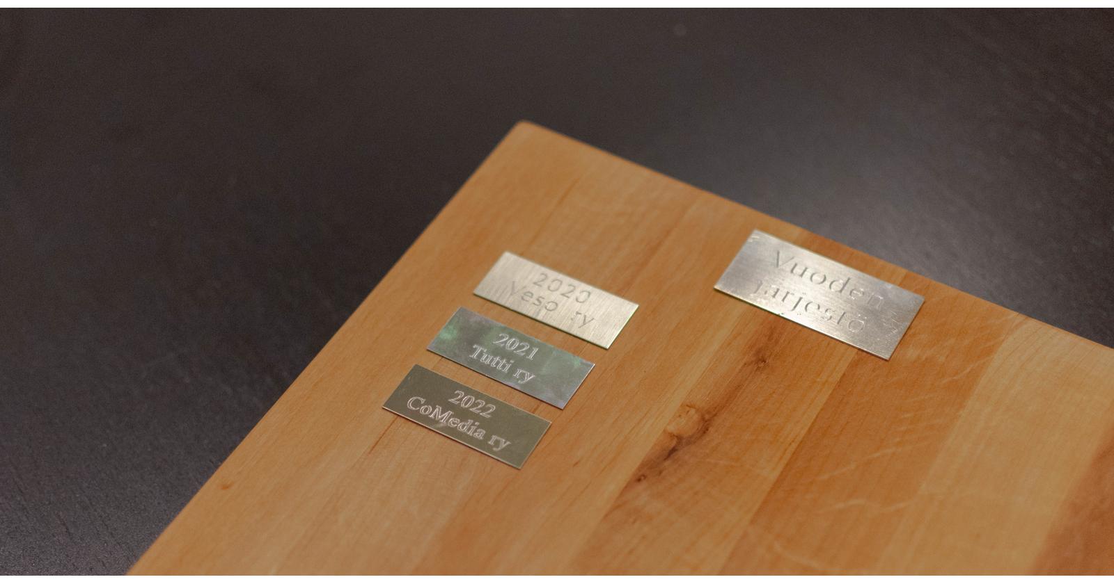 Puisella laudalla neljä pientä metallista kylttiä, jossa yhdessä luee CoMedia ry 2022