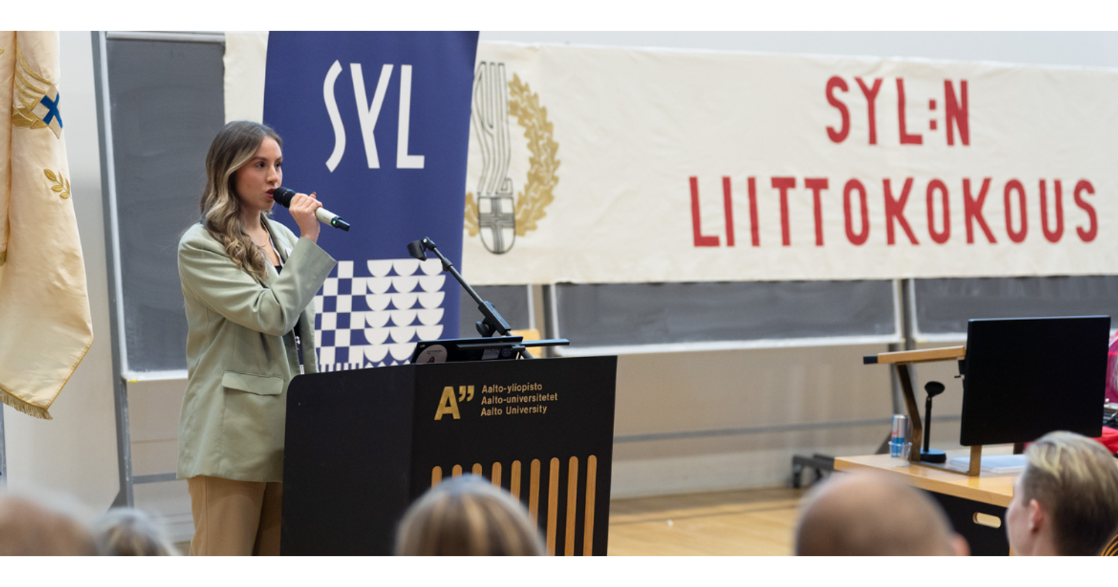 Maria Saita speaking into the microphone in the speaker's booth. There is an Aalto University logo on the speaker's booth and in the background a Rollup with the SYL logo and a banner with the words SYL General Assembly on it. In the foreground of the picture you can see people sitting in the audience.