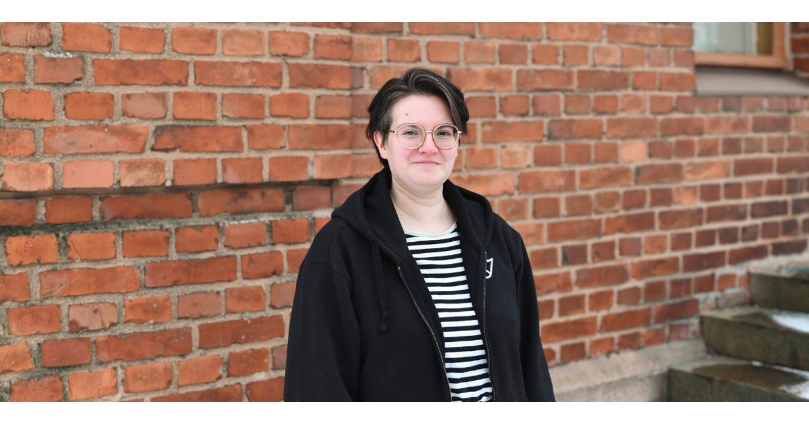 Aino Hyyryläinen stands in front of a red brick wall. They are smiling at the camera and wearing a black hoodie with VYY's logo.