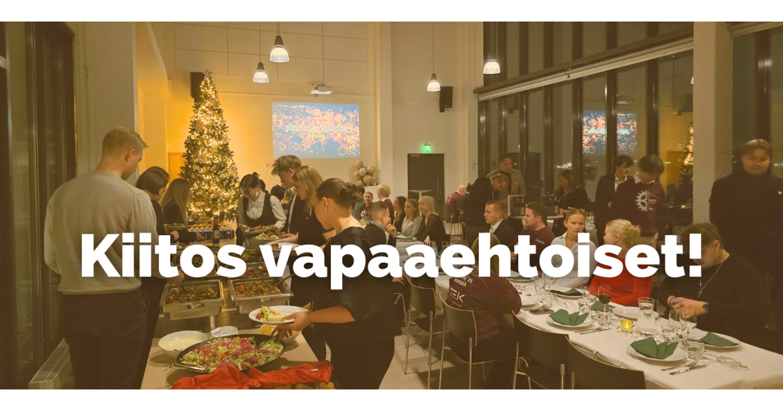 Valokuvassa ihmisiä yhteisessä juhlassa, osa istuu pöydissä ja osa hakee seisovasta pöydästä ruokaa. Taustalla näkyy koristeltu joulukuusi. Kuvan päällä on teksti Kiitos vapaaehtoiset!