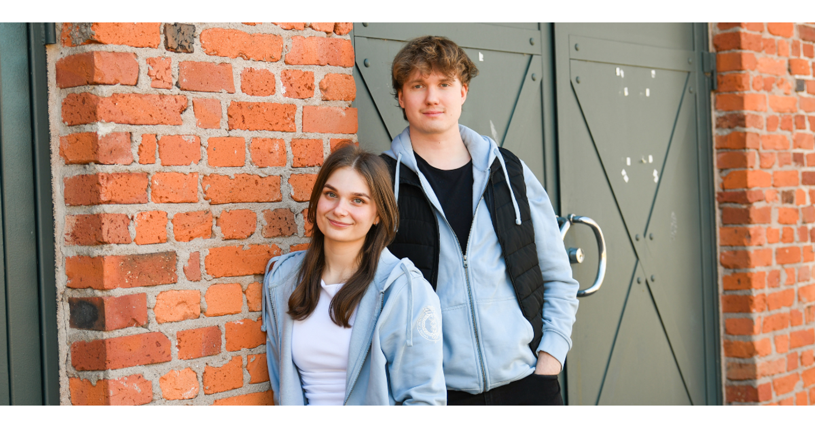 Valokuvassa Saana Sammalo ja Aleksi Lumme hymyilev&auml;t kameralle ja nojaavat tiilisein&auml;&auml;n.