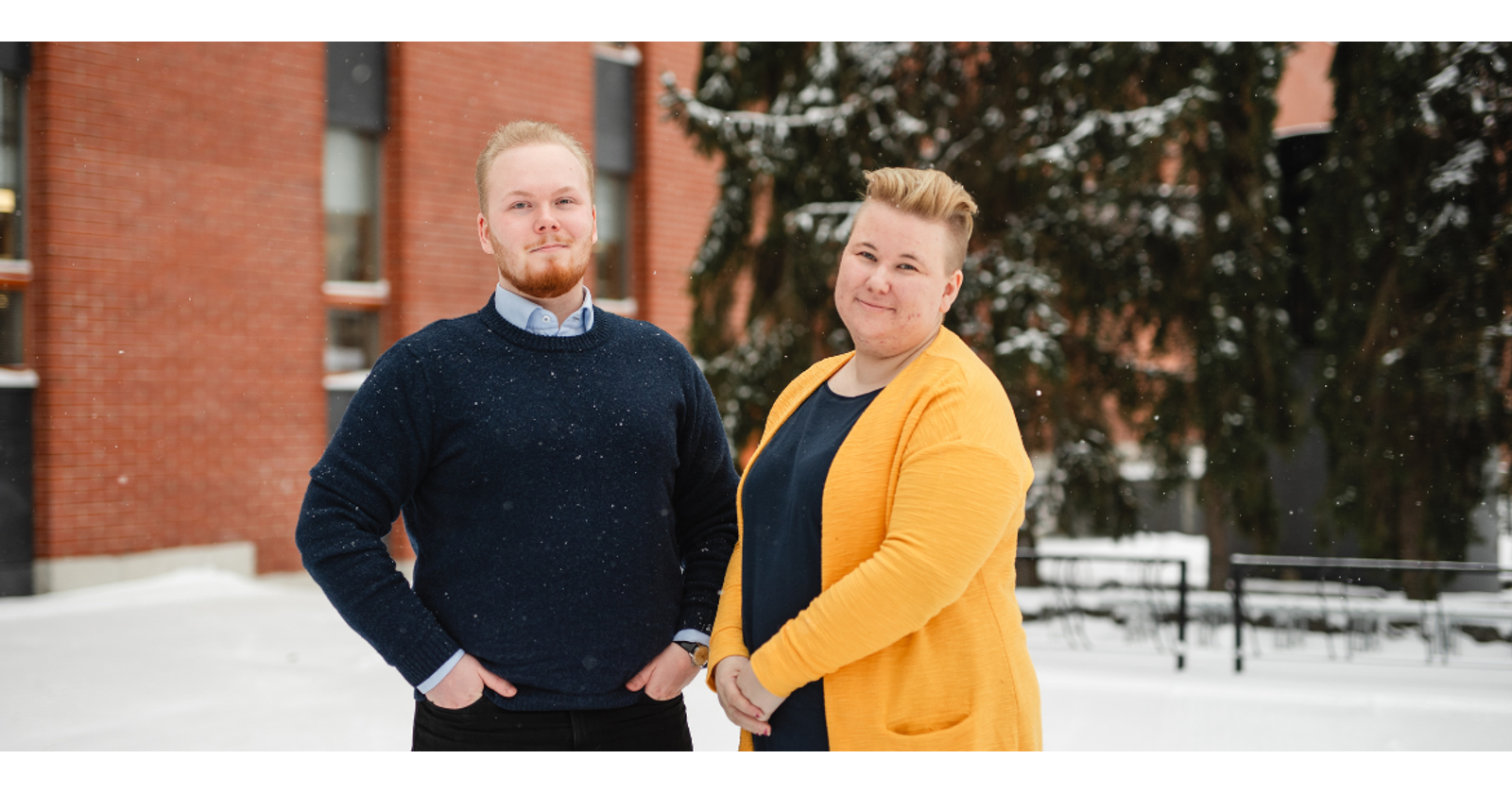 Juuso Tynjälä ja Maaria Salminen seisovat Vaasan yliopiston kampuksen edustalla. Taustalla on tiiliseinä ja havupuita. Maassa on lunta ja kuvan etualalla leijailee lumihiutaleita. Juuso ja Maaria katsovat kameraan ja hymyilevät.