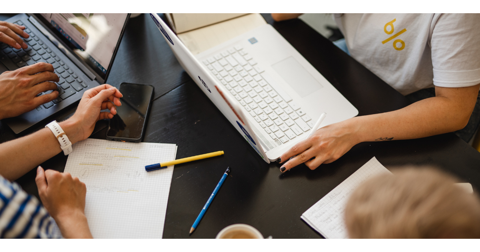 Overhead photo of people working around a table. The photo shows arms and hands, laptops, written notes, and pens.