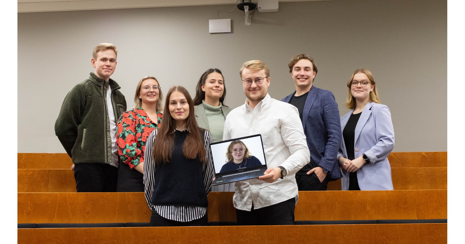 Seitsemän hallituslaista seisoo luentosalissa rivissä ja yhdellä on kannettava tietokone kädessään