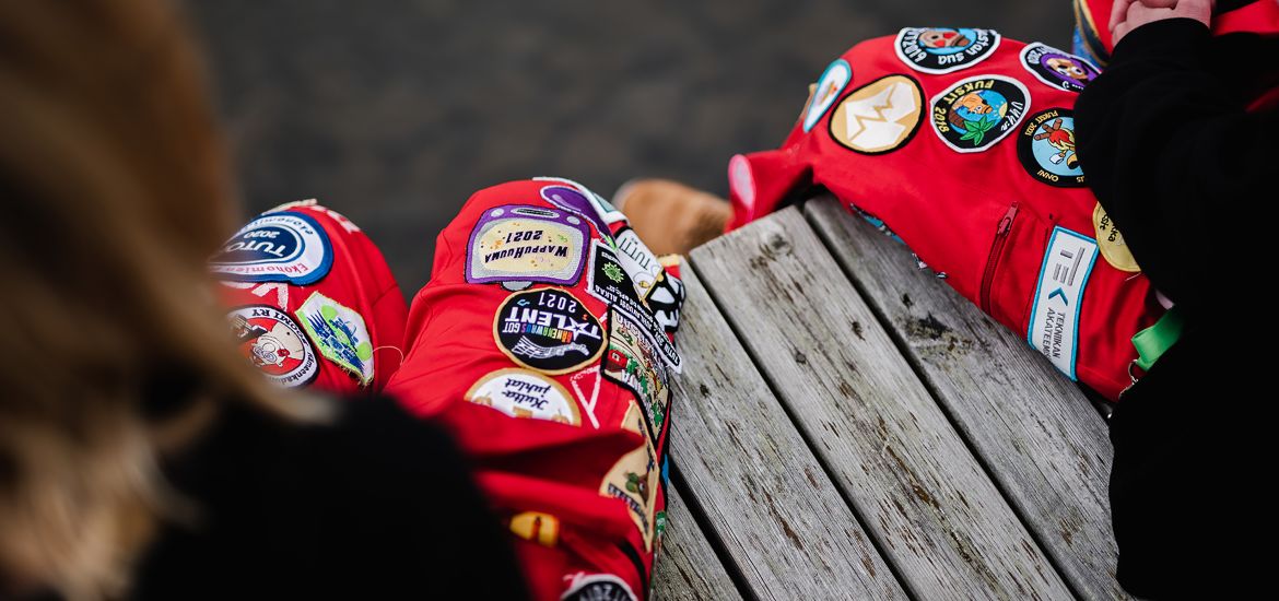 Two people are sitting on a dock wearing red overalls and dark tops. There is dark water in the background. The image is taken from the shoulder level of the people in the picture towards the overalls and the dock.